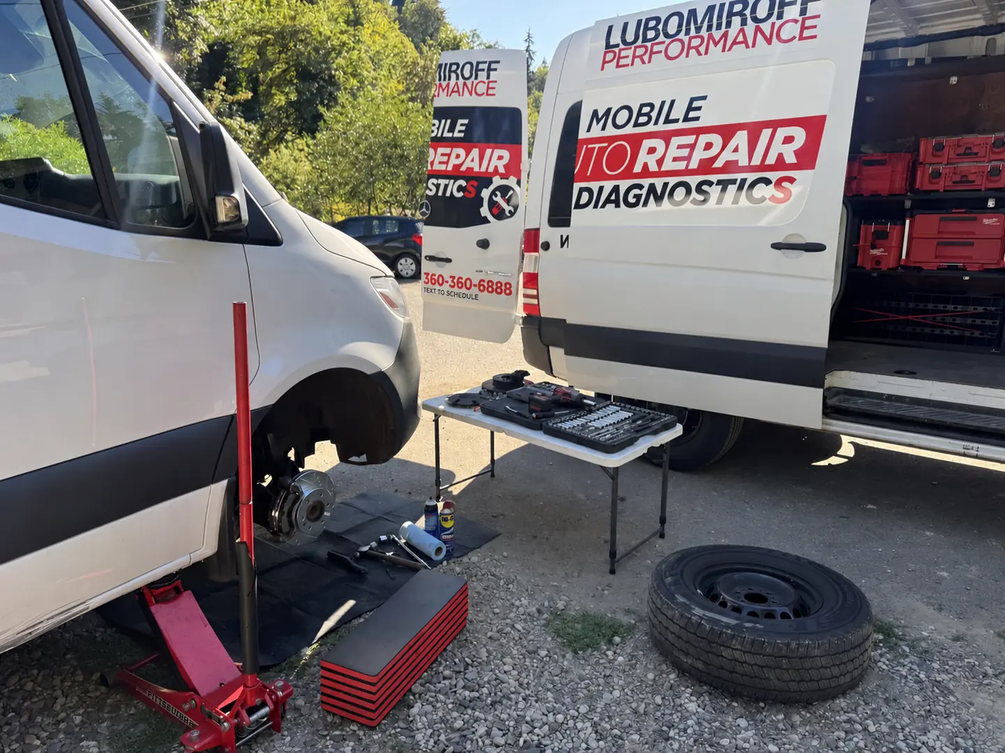 White Mercedes-Benz Sprinter with a front wheel removed beside the Lubomiroff Performance service van during mobile brake service.