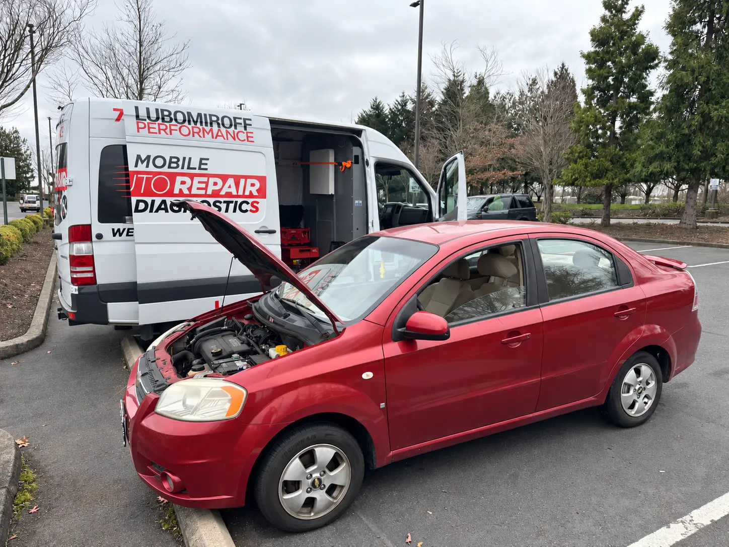 Red Chevrolet Aveo with its hood open beside the Lubomiroff Performance service van.