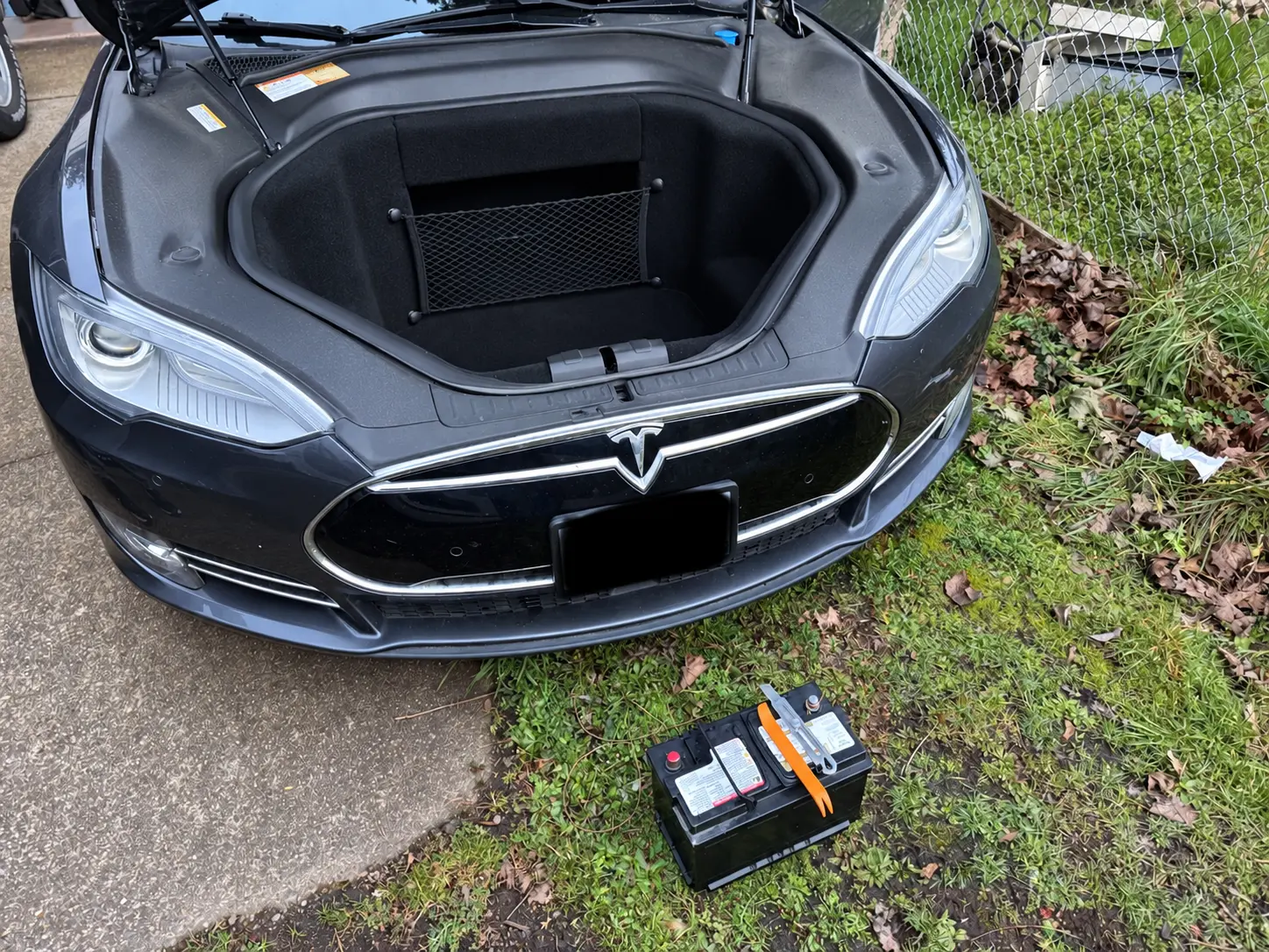 Tesla Model S with the front trunk open and a low-voltage battery on the ground in front.
