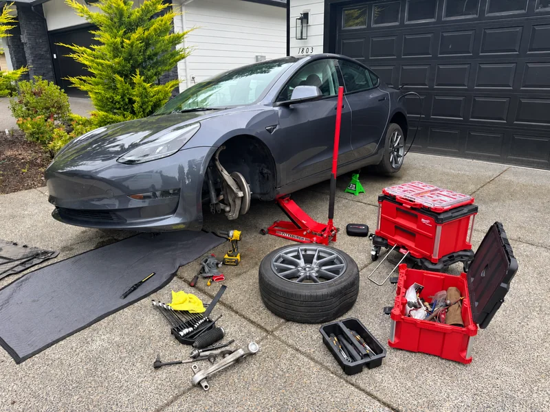 Gray Tesla Model 3 raised with a front wheel removed and suspension tools beside it